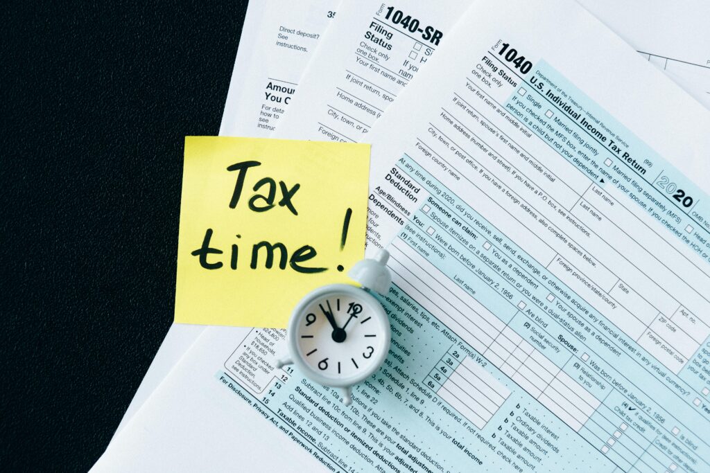 pexels photo 6863259 6863259 A stack of tax forms with a clock and yellow sticky note saying 'Tax time!' indicating urgency.